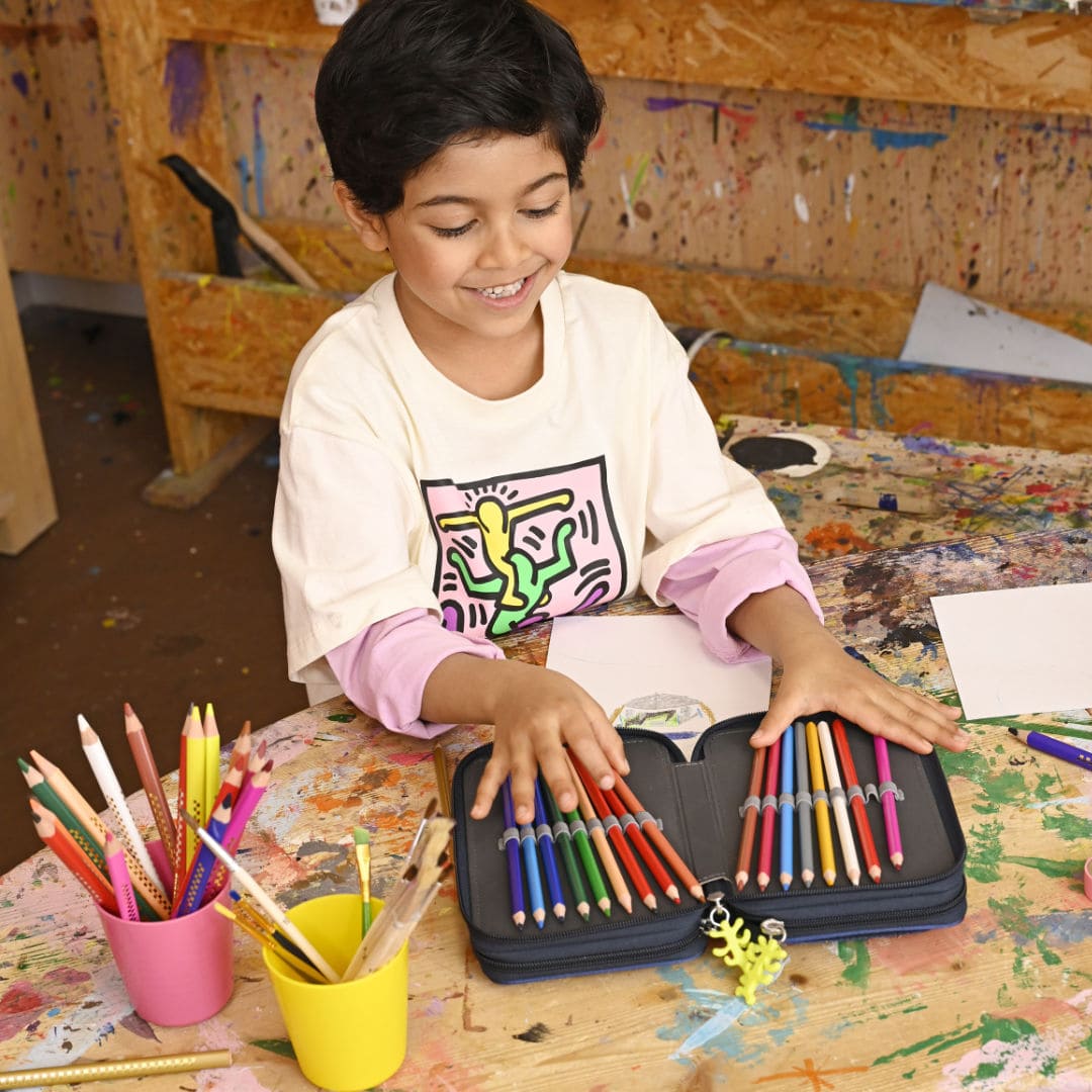 ein Schuljunge zeigt auf einem Kunsttisch aus Holz die Buntstifte in seinem geoeffneten Schulmaeppchen | KoenigPlus