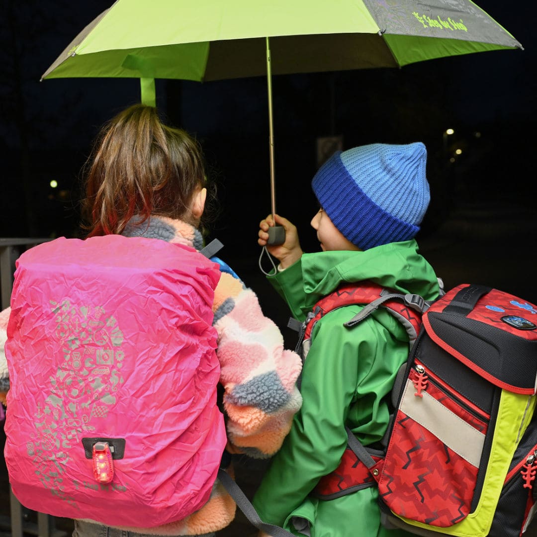 eine Schueler mit Ranzen und pinkem Ranzen-Ueberzieher zusammen mit einem Schueler mit neongelbem Pullover an rotem Ranzen, beide von hinten unter einem Regenschirm bei Dunkelheit | KoenigPlus