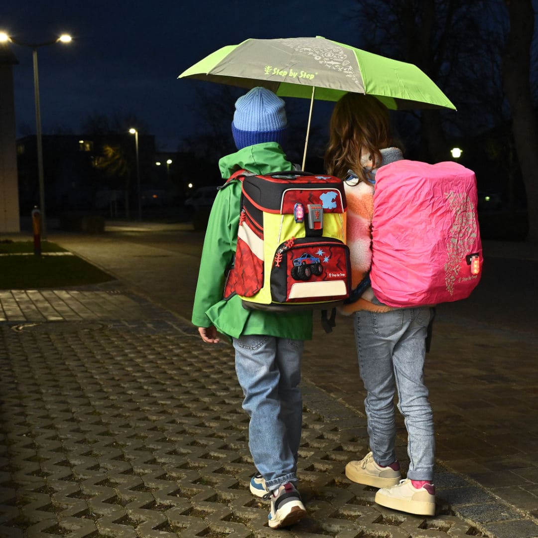 zwei Schueler gehen mit geoeffnetem Regenschirm in Dunkelheit auf Strasse. Der eine Ranzen ist mit einem Ueberzieher geschuetzt, der andere durch einen Pullover | KoenigPlus