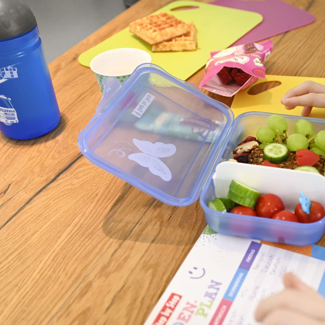 angeschnitten eine blaue Kunststoff-Trinkflasche, eine geoeffnete, Obstgefuellte Lunchbox aus Kunststoff in blau und ein Stundenplan auf einem Holztisch | KoenigPlus