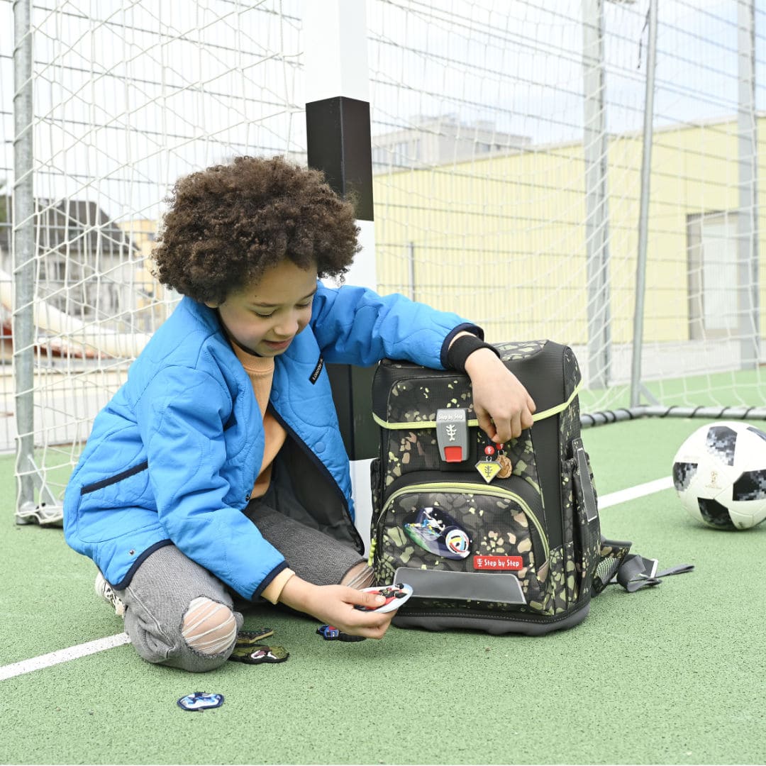Schueler sitzt vor Fussballtor mit Ranzen und sucht aus vier Step by Step MagicMags das passende Dekorelement aus.|KönigPlus