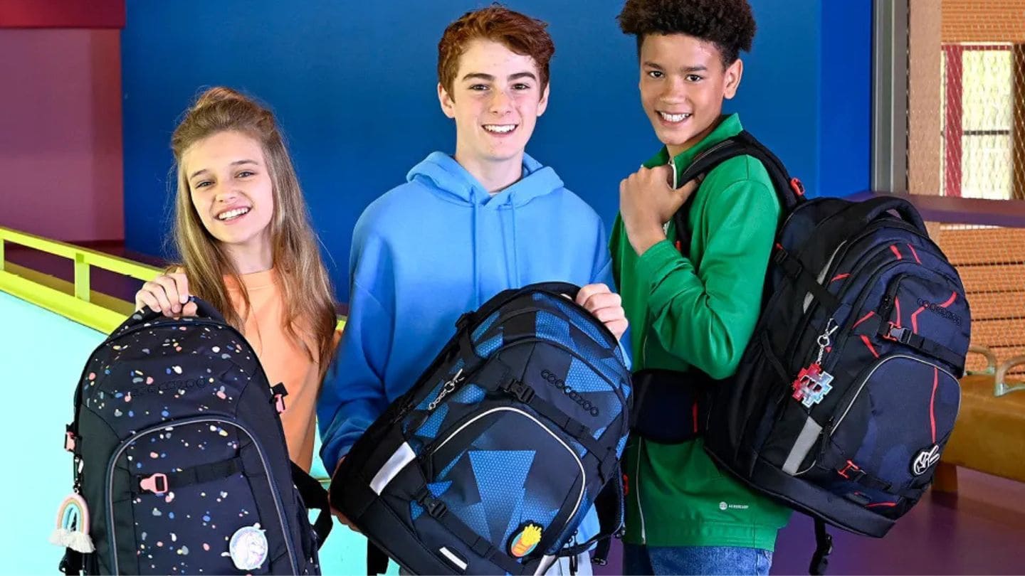 freundlich schauende ein Girl und zwei Boys zeigen ihre Rucksaecke in einem bunten Schultreppenhaus| KoenigPlus