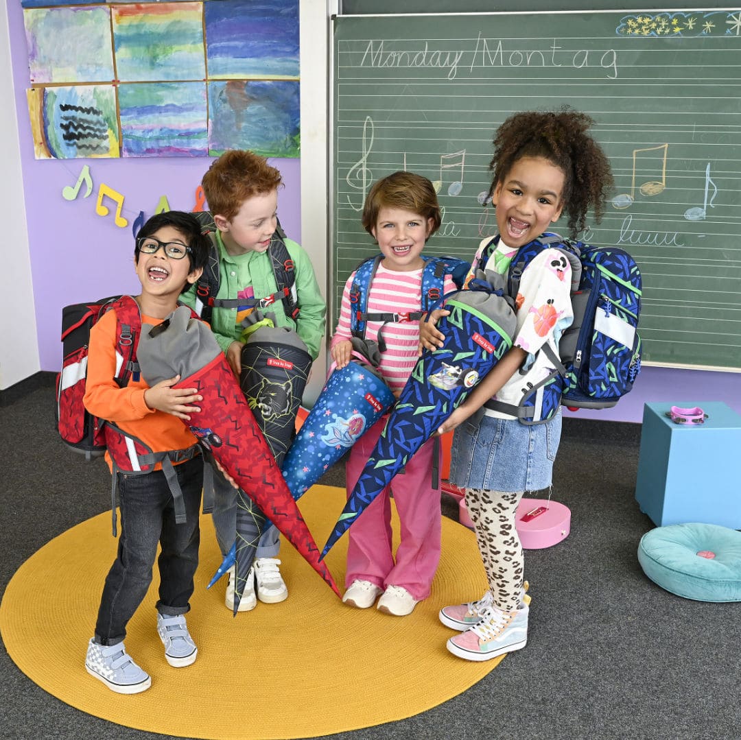 froehliche zwei Schuelerinnen und zwei Schueler mit Ranzen auf dem Ruecken und Schultueten vor sich im Klassenzimmer vor der Tafel | KönigPlus