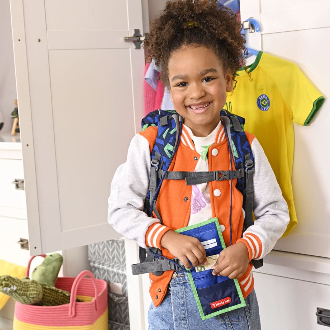 laechelnde Grundschuelerin mit Rucksack auf dem Ruecken und umgehaengten Brustbeutel vor dem geoeffneten Kleiderschrank im Kinderzimmer| KoenigPlus